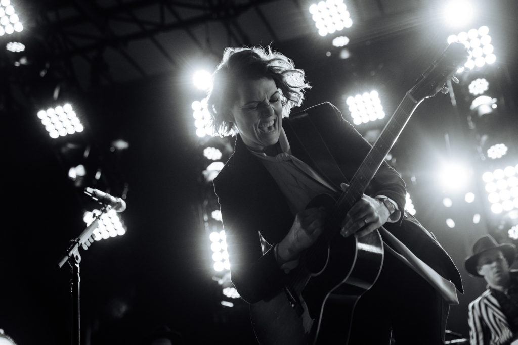 Brandi Carlile with hair flowing and a giant grin as she plays her guitar in motion