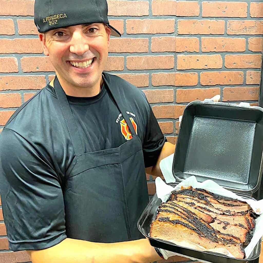 Man holding a plate of brisket. He's wearing a black hat and a black apron.