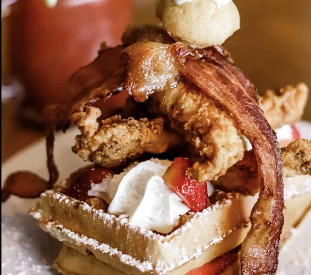 A waffle covered in powdered sugar and topped with fried chicken, bacon, and fresh fruit