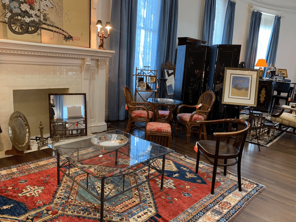 a drawing room filled with antique furniture. There's a fireplace on the left side with three windows on the far wall covered by blue curtains. 