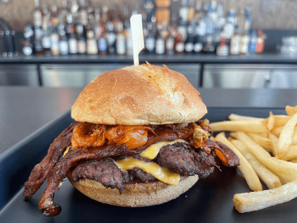 A burger covered in fresh bacon and kimchi. The burger is on a toasted bun on a black plate with a side of golden fries. 