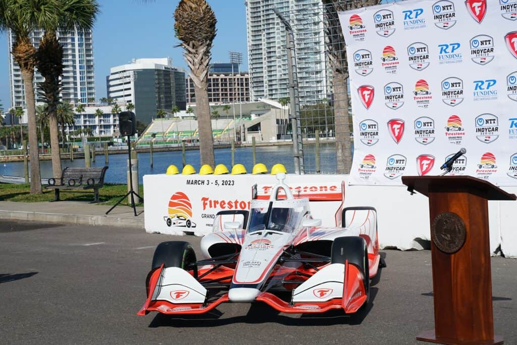 An indycar parked on the waterfront. A podium is place to the side of the car with a large step and repeat with Grand Prix branding behind the car. 