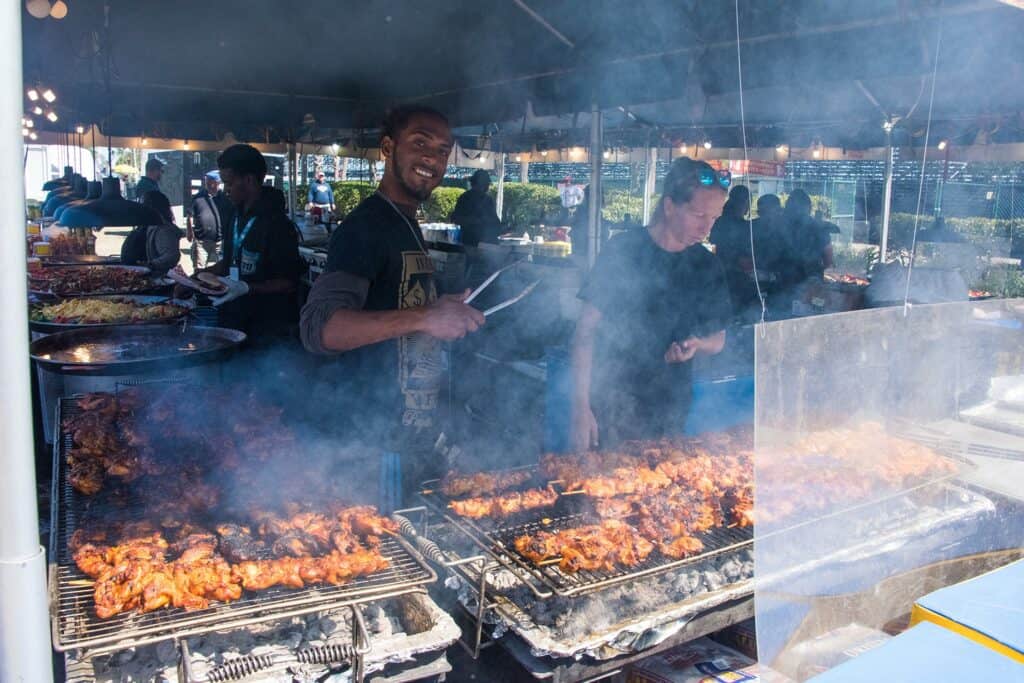 Someone behind a grill with chicken smoking. The chef is holding a pair of tongs and smiling. 