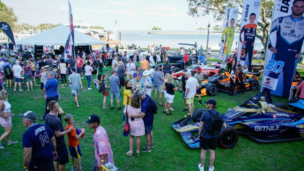 Large gathering in a waterfront park. Race cars are arranged on a lawn. 