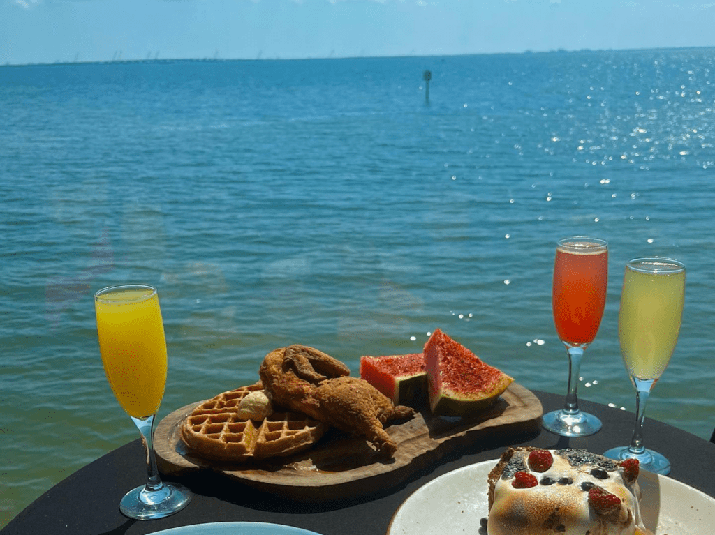 a plate of waffles and chicken with watermelon on the side with several flutes filled with mimosas. The plate is set on a table with a waterfront view. 